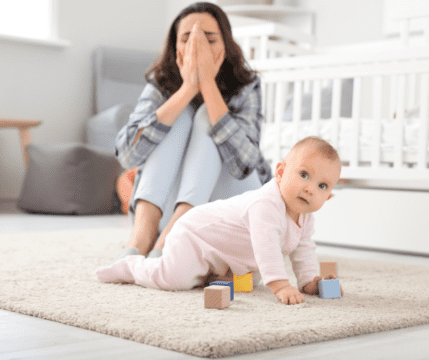 kobieta siedzi na dywanie, zakrywa ze smutki rękami twarz, obok niej raczkujące niemowlę bawi się klockami